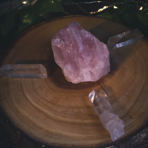 a simple crystal grid set up on a slice of wood. a large rose quartz sits in the center with 3 clear quartz radiating out from the center stone.